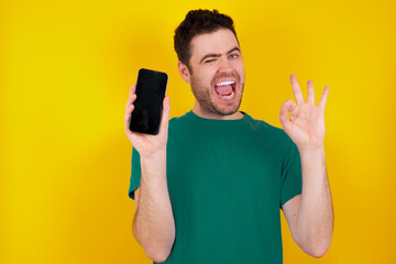 young handsome caucasian man wearing green t-shirt against yellow background holding in hands cell...