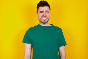 Portrait of dissatisfied young handsome caucasian man wearing green t-shirt against yellow wall smirks face, purses lips and looks with annoyance at camera, discontent hearing something unpleasant