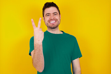 young handsome caucasian man wearing green t-shirt against yellow background smiling and looking friendly, showing number two or second with hand forward, counting down