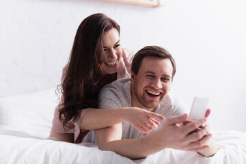 Cheerful woman pointing with finger at smartphone on blurred foreground near husband in bedroom