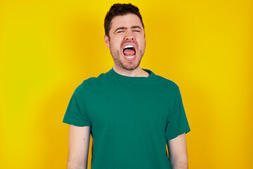Fototapeta premium young handsome caucasian man wearing green t-shirt against yellow background yawns with opened mouth stands. Daily morning routine