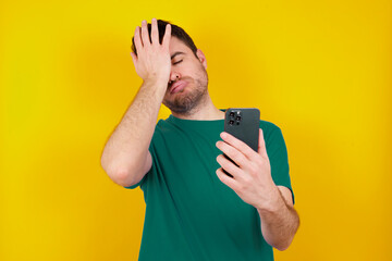 Upset depressed  young handsome caucasian man wearing green t-shirt against yellow background makes face palm as forgot about something important holds mobile phone expresses sorrow and regret blames