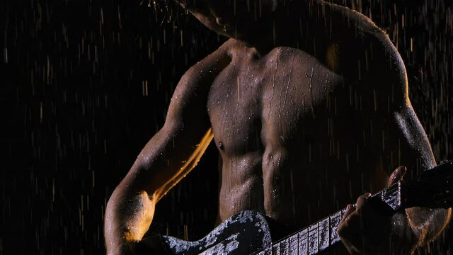 A man with a naked torso, wet from raindrops, plays the guitar while sitting on the surface of the water. The musician plays the guitar in the rain, romance, night. Close up. Slow motion.