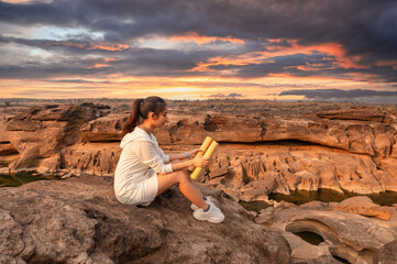 Beautiful young asian woman traveler sitting and looking at paper map on rock cliff in grand canyon of Thailand