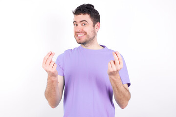 young handsome caucasian man wearing purple t-shirt against white background keeps palms together, has pleased expression. Glad attractive male makes request, pleads for mercy. Hopeful young adult.
