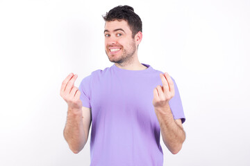 Naklejka premium young handsome caucasian man wearing purple t-shirt against white background keeps palms together, has pleased expression. Glad attractive male makes request, pleads for mercy. Hopeful young adult.