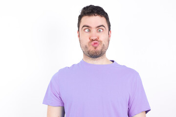 Fototapeta premium young handsome caucasian man wearing purple t-shirt against white background crosses eyes, puts lips, makes grimace with awkward expression has fun alone, plays fool.