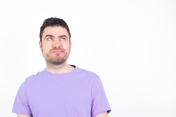 young handsome caucasian man wearing purple t-shirt against white background has worried face looking up lips together, being upset thinking about something important, keeps hands down.