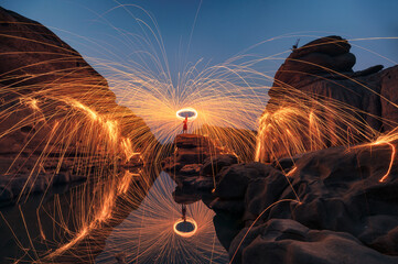 Man wielding rounded burning fire spark from steel wool in rock rapids of grand canyon reflection...