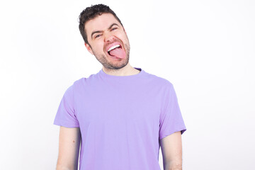 Fototapeta premium young handsome caucasian man wearing purple t-shirt against white background with happy and funny face smiling and showing tongue.