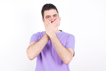 Upset young handsome caucasian man wearing purple t-shirt against white background, covering her mouth with both palms to prevent screaming sound, after seeing or hearing something bad.