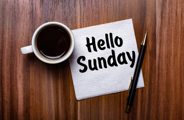 On a wooden table next to a white cup of coffee and a pen is a white paper napkin with the words HELLO SUNDAY