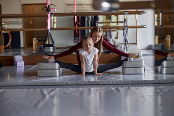 Young confident female teacher is helping girl with stretching in modern hall