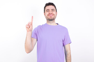 Naklejka premium young handsome caucasian man wearing purple t-shirt against white background showing and pointing up with fingers number one while smiling confident and happy.