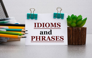 IDIOMS and PHRASES - words on a white sheet with clamps against the background of a cactus and jars with pencils - obrazy, fototapety, plakaty