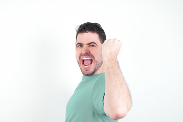 Overjoyed young handsome caucasian man wearing green t-shirt against white background glad to receive good news, clenching fist and making winning gesture.