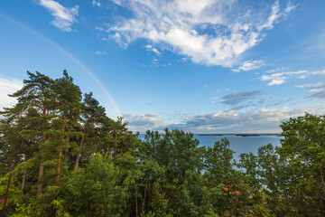 Beautiful view of tops of green trees on blue sky with a rainbow after rain on background Baltic sea. Sweden.