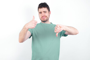 Fototapeta premium young handsome caucasian man wearing green t-shirt against white background feeling unsure making good bad sign. Displeased and unimpressed.