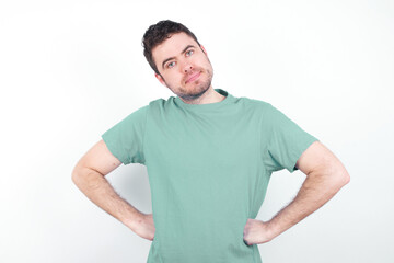 Obraz premium Funny frustrated young handsome caucasian man wearing green t-shirt against white background holding hands on waist and silly looking at awkward situation.