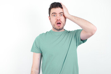 Portrait of confused young handsome caucasian man wearing green t-shirt against white wall holding hand on hair and frowning, panicking, losing memory. Worried and anxious can not remember anything.