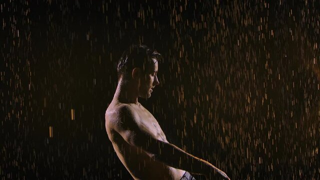 A Wet Man With A Naked Torso Moves Sexually, Breaking Raindrops. Silhouette Of A Male Playing The Guitar On A Black Background Under The Streams Of Rain With Studio Light. Close Up. Slow Motion.