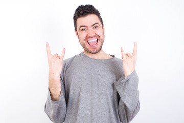 Fototapeta premium young handsome caucasian bearded man wearing pyjama over white wall makes rock n roll sign looks self confident and cheerful enjoys cool music at party. Body language concept.