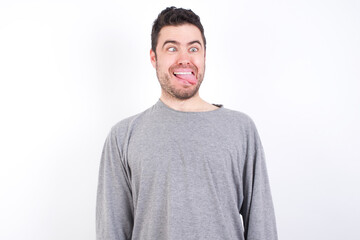 Funny young handsome caucasian bearded man wearing pyjama over white wall makes grimace and crosses eyes plays fool has fun alone sticks out tongue.