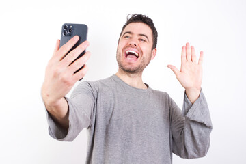 young handsome caucasian bearded man wearing pyjama over white wall holds modern mobile phone and makes video call waves palm in hello gesture. People modern technology concept