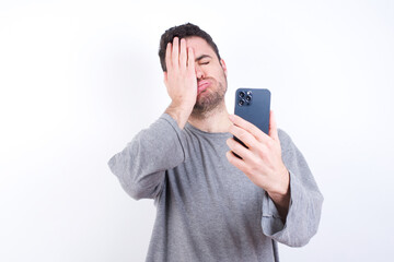 Upset depressed young handsome caucasian bearded man wearing pyjama over white wall makes face palm as forgot about something important holds mobile phone expresses sorrow and regret blames