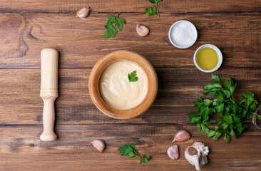 Homemade aioli sauce in a wooden mortar on a table with the mallet on the side, the garlic, olive oil, parsley and salt
