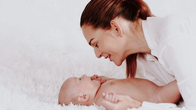 Attractive young mother with tail in white shirt kissing her newborn baby laying on soft white background. Copy space