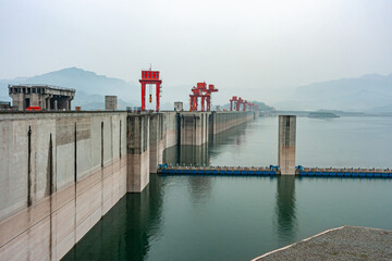 Three Gorges Dam, China - May 6, 2010: Yangtze River. Up-river shot of massive and long beige concrete dam with red flood control door lifts on top under foggy morning sky and green water up front. 