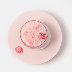 Rose-scented beauty sea salt in jar with cap and rose flowers on round podium. Cosmetic product top view, pink pastel colored