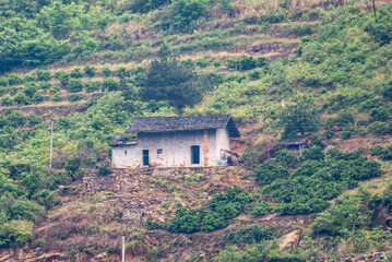 Fototapeta na wymiar Three Gorges Dam, China - May 6, 2010: Closeup of small white farm building with dark gray roof set on green hill slope with terraced agriculture. Patches of brown dirt.