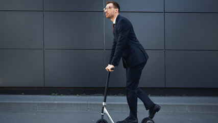 full length of businessman in formal wear and glasses riding e-scooter near building.