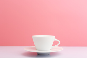 White ceramic coffee cup with saucer on white table against light pink background with copy space