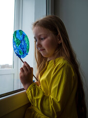 Fototapeta premium Cute little girl painting planet on window at home. Happy Earth Day April 22 greeting message. Creative family leisure lockdown new reality. Ecology Saving environment conscious consumption concept.