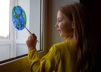 Cute little girl painting planet on window at home. Happy Earth Day April 22 greeting message. Creative family leisure lockdown new reality. Ecology Saving environment conscious consumption concept.