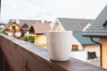 The steam rises from a cup of hot coffee or tea on the balcony with mountains and roofs of houses on the background. 