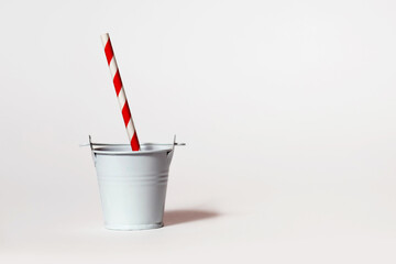 One white toy bucket on a white background with a striped red and white drinking straw. Weekend party concept with non-standard serving drinks or thirst. Free space for text