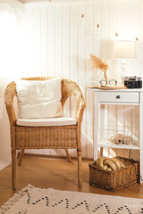 Wicker peacock chair with pillow and bedside table with book, retro camera, glasses and lamp in beige light wooden interior
