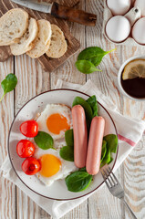 Breakfast served on a light wooden table. Fried eggs with sausages, spinach and cherry tomatoes, on a round plate