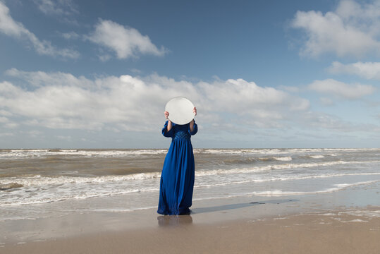woman  in long blue vintage dress standing on the beach near the sea covering her face with a round mirror  reflecting the clouds and sky