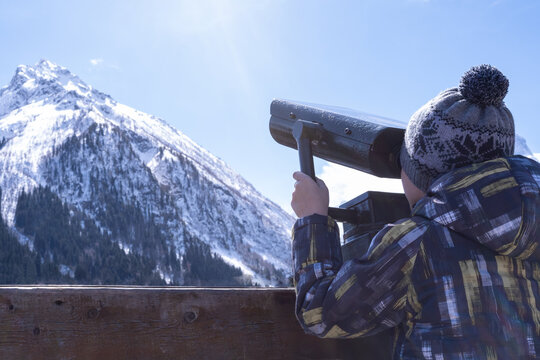 A child in a bubon hat looks through binoculars into the distance at a beautiful snowy mountain top in a bright sunny sky