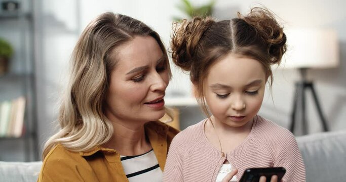 Close-up of beautiful mother helping her little daughter to understand how to use smartphone and searching cartoons in Internet.
