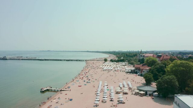 Aerial view of a large sandy beach with white umbrellas and people sunbathing on the Baltic Sea in Sopot, Poland. Cinematic drone shot on a sandy beach with crowds of tourists swimming and relaxing