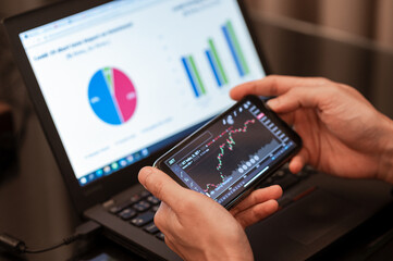 Close up hands of businessman analyzing financial data on smartphone and computer screen background.