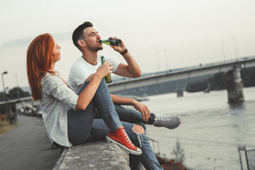 Young couple sitting on dock by the river ,relaxing and drinks beer.	
