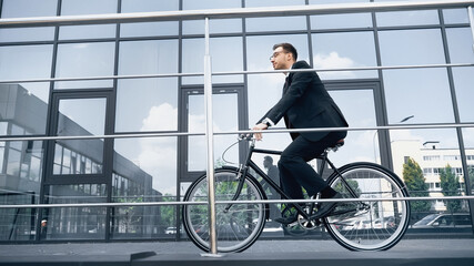 full length of businessman in suit and glasses riding bicycle near building with glass facade.