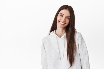 Beautiful young woman with long dark hair, tilt head and smile at camera tenderly, standing candid and positive against white background, wearing hoodie